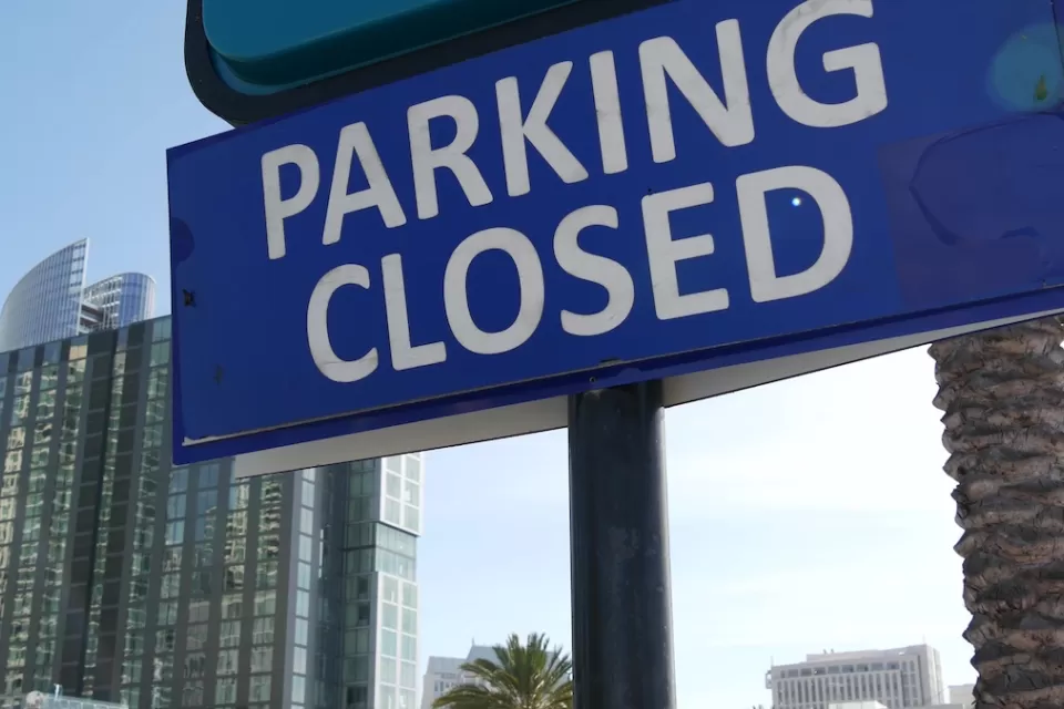 Close-up of blue sign with white text reading PARKING CLOSED with urban glass buildings in background.